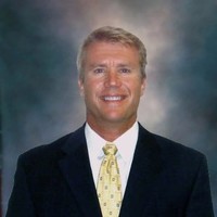 Ryan Zenk in a dark suit and yellow tie stands in front of a blurred, neutral background, smiling at the camera.
