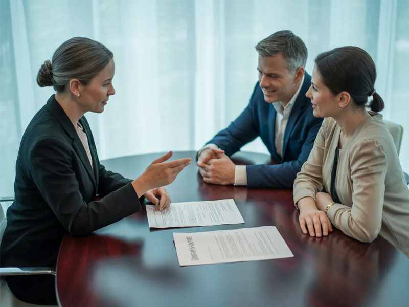 Three people sit at a round table with documents, engaged in a business discussion. One woman gestures while the other two, a man and woman, listen attentively.